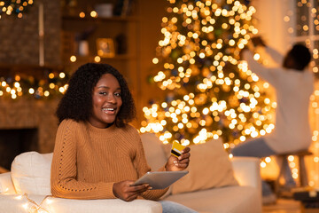 Smiling young african american lady sits comfortably on a couch, using a tablet and credit card for online shopping. In the background, a man decorates a Christmas tree with lights.