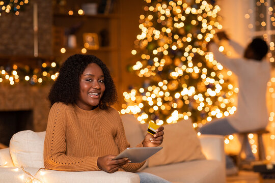Smiling young african american lady sits comfortably on a couch, using a tablet and credit card for online shopping. In the background, a man decorates a Christmas tree with lights.