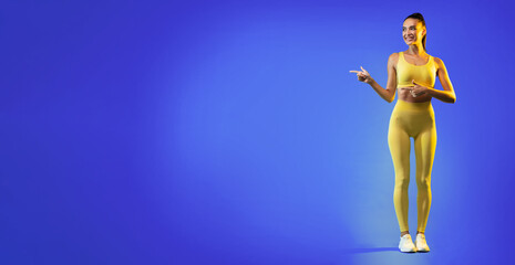A fitness instructor stands confidently on a blue background, wearing bright yellow activewear. She gestures playfully, inviting engagement during a workout session.