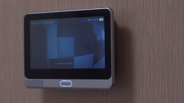 A smart video peephole on a door rings, its monitor lighting up to show the outside view. The device makes a simple, high-pitched ding sound.