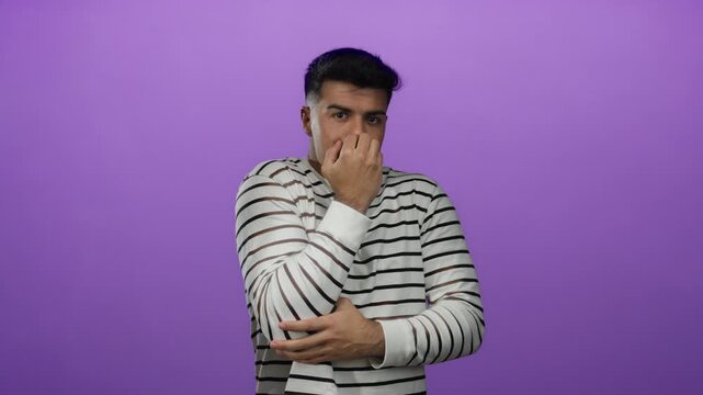 Young hispanic man looks anxious in striped shirt against isolated purple background.