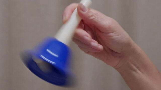 A hand rings a blue, metal handbell with a white handle. The bell produces a clear, high-pitched ringing sound.