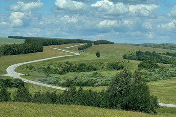 A winding asphalt road winds through green hills and meadows, with clusters of trees and shrubs along the edges, and a blue sky with fluffy clouds above the landscape.