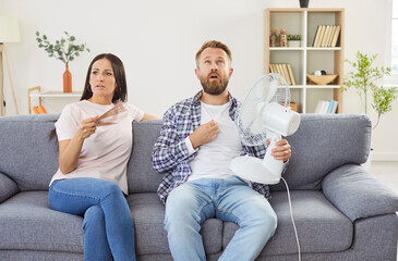 Young family couple sitting on sofa at home suffering from awful summer heat wave. Sweaty exhausted man and woman trying to cool themselves with electric fan and get some fresh air in overheated room.