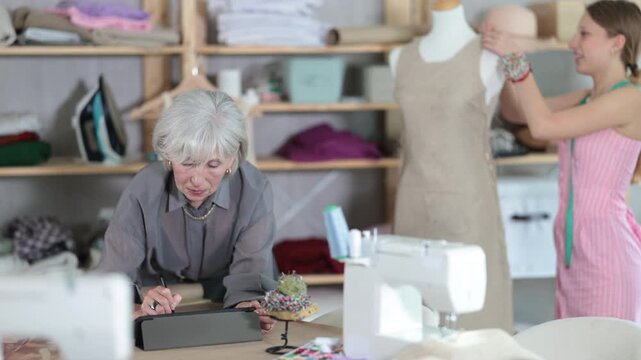 Positive mature tailoress drawing dress model in tablet while other working with mannequin in background. High quality 4k footage
