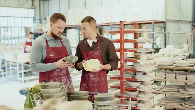Two enthusiastic young male potters in stained burgundy aprons examining handcrafted ceramic wares in studio and exchanging ideas about techniques of casting