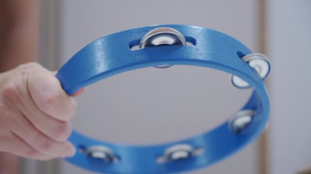 A hand shakes a plastic tambourine with metal jingles. The percussion instrument creates a loud, rhythmic metallic jingle.