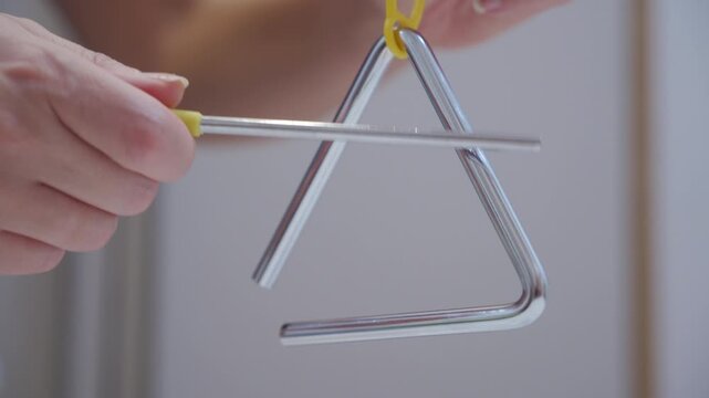 A hand strikes a metal triangle with a metal beater. The percussion instrument produces a high-pitched, sustained metallic ringing tone.