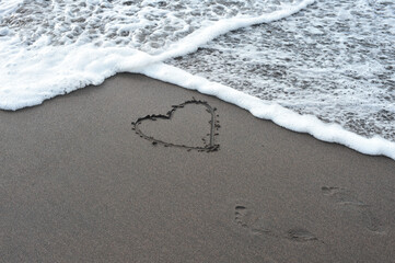 Heart drawn in wet sand on tropical beach with ocean wave, foam, and footprints. Concept of love, romance, vacation, and ephemeral beauty.