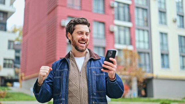 Joyful man stands outside, excitedly looking at phone. Good news brings triumphant smile and fist pump gesture. Happiness and success radiating from the scene.