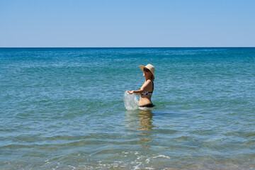 Happy middle-aged woman in a hat splashing water in the sea, enjoying her vacation, the sun