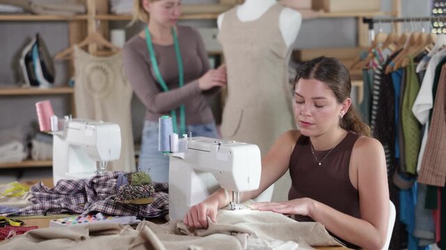 Female tailor is working on a sewing machine, checking the neatness of the stitches against the background of an assistant and a mannequin. Two tailors in a workshop