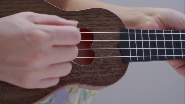 A hand strums the nylon strings of a wooden ukulele with a plastic pick. The stringed instrument emits a full, light chord strum.