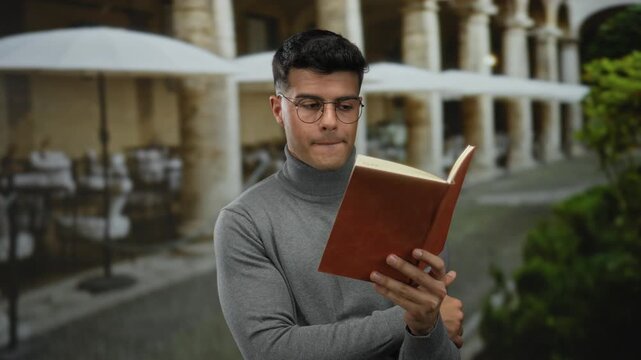 Young man in glasses reads a book on a terrace at an outdoor city restaurant, surrounded by greenery and umbrellas, providing a serene and studious ambiance.