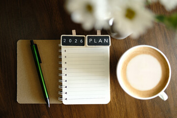 A '2026 PLAN' blackboard clip on a notebook, pen, coffee, and flowers, laid flat on a brown wooden table. Concepts of business planning, strategy, financial goals, and New Year resolutions.
