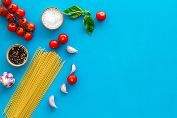 Flat lay of uncooked spaghetti with ingredients for cooking Italian pasta - tomatoes and basil with garlic - on a blue background, top view