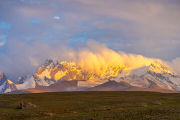 Panoramic view of a range of snowcapped mountains at sunset or sunrise, with their slopes glowing bright gold from the light breaking through low clouds over a wide, dark foreground plain