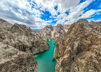 A very high angle, wide view of a turquoise glacial lake squeezed between immense, rugged, textured mountain rock formations under a cloudy blue sky