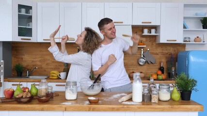 Energetic young couple having fun while preparing breakfast in their modern kitchen, singing into wooden spoons as microphones and dancing joyfully together in a bright, happy home setting - Powered by Adobe