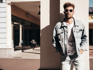 Portrait of handsome confident stylish hipster lambersexual model. Sexy man dressed in jeans jacket. Fashion male posing in the street. Taking off sunglasses in Europe city. Outdoors at sunset