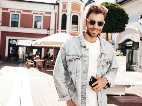Portrait of handsome smiling stylish hipster model. Sexy man dressed in jeans jacket. Fashion male posing in the street. In sunglasses in Europe city. Outdoors at sunset