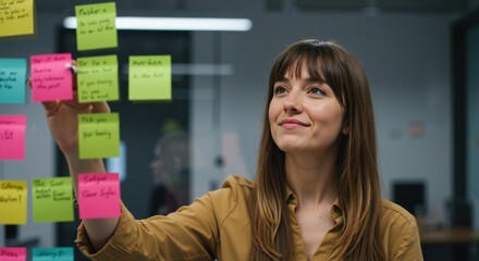 Creative businesswoman brainstorming with sticky notes on glass wall. Young professional planning project strategy in modern office. Agile methodology and task organization concept