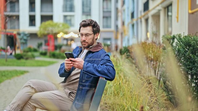Man sitting on park bench, smiling while interacting with phone. Expresses contentment and happiness through digital connection. Casual outdoor moment.
