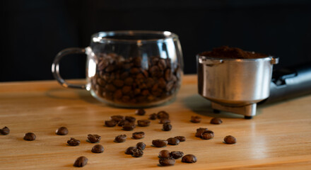 Close-up of roasted coffee beans scattered on a wooden table beside a glass cup and espresso portafilter, symbolizing aroma and coffee preparation.