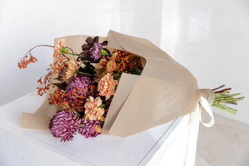 Lush autumn floral bouquet featuring purple chrysanthemums, peach carnations, and vibrant orange rowan berries wrapped in natural kraft paper on a white surface