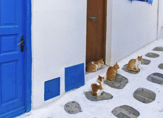 Narrow alleyway in Mykonos with white-painted ground and walls, accented by bright blue doors and window frames, featuring four orange and white cats