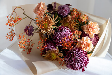 Vibrant autumn bouquet featuring dark purple chrysanthemums, delicate peach carnations, and bright orange berries, wrapped in rustic brown paper on a sunlit white table