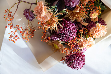 Vibrant autumn bouquet featuring dark purple chrysanthemums, delicate peach carnations, and bright orange berries, wrapped in rustic brown paper on a sunlit white table