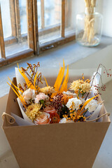 Autumnal floral arrangement featuring peach roses, yellow chrysanthemums, protea, and orange accents, neatly packaged in a brown craft box by a window