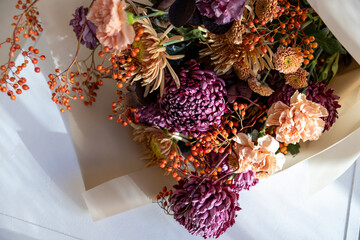 Vibrant autumn bouquet featuring dark purple chrysanthemums, delicate peach carnations, and bright orange berries, wrapped in rustic brown paper on a sunlit white table