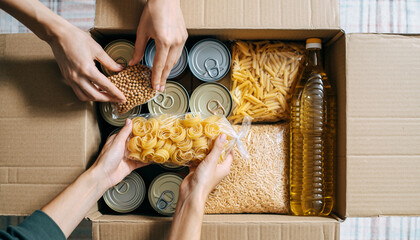 Hands placing canned food pasta grains and cooking oil into cardboard donation box top view concept of charity food drive and community support warm and caring atmosphere