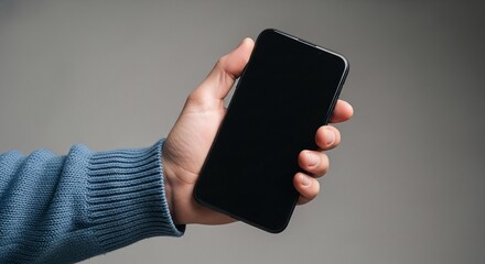 A person holding a black smartphone in their hand wearing a blue sweater against a gray background