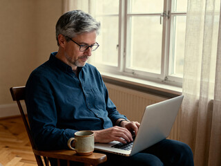 A man is focused on his laptop in a serene, softly lit room while enjoying a coffee. Freelance lifestyle aesthetic