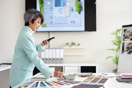  Concentrated  interior designer womanwith blueprint drawing ,swatches and material , discussing interior design with client  by phone, choosing materials for decorations.