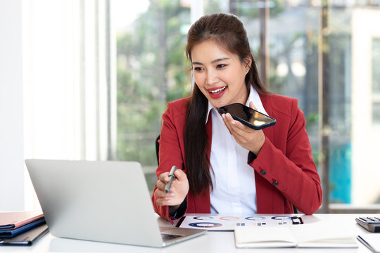 Smiling  businesswoman in modern office , holding smartphone recording voice message or lead conversation on speakerphone. Business, remote communication, voice recognition, AI - Powered by Adobe