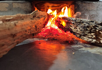 Naklejka premium Burning firewood inside a rustic fireplace with warm orange flames and glowing embers. Perfect for concepts of warmth, comfort, coziness, rustic lifestyle, and winter evenings.