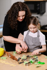 Mother helps daughter cut cookie dough with cutters at kitchen table. Family baking activity in kitchen, where mom teaches daughter art of baking.