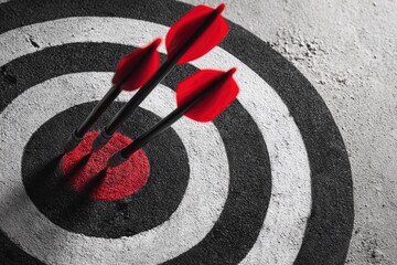 A close-up view of a bullseye target painted with concentric black and white circles on a rough, textured surface, possibly concrete or asphalt. Three red-fletched darts are perfectly embedded in the 