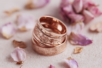 Elegant rose gold wedding rings with delicate floral design surrounded by soft pink petals on textured surface, perfect for romantic celebrations.