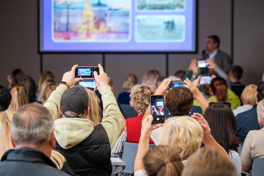 Crowd watches speaker on stage at conference while attendees record slides with smartphones and cameras creating dynamic social video moment