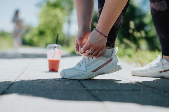 A close-up of a woman adjusting her athletic shoes in a park setting, highlighting fitness, outdoor activity, and hydration with a smoothie in focus.