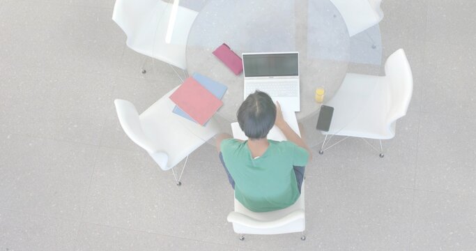 Studying Asian student in green T-shirt using laptop at round glass table, with notebooks and drink