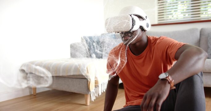 Sitting man wearing white VR headset and orange tee on wood floor near sofa, holographic mist