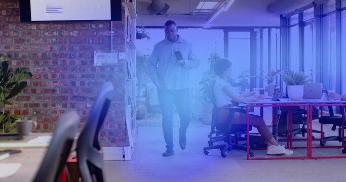 Walking colleague in light shirt and dark pants checking smartphone in modern office near red desks - Powered by Adobe