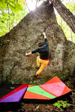 A rock climber climbs the face of a boulder with crash pads below