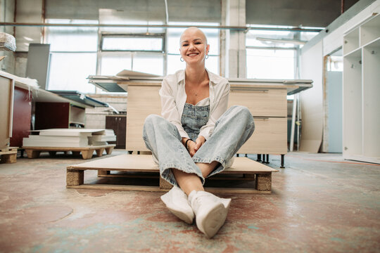 Bald woman smiling in overalls sitting on pallet at furniture workshop